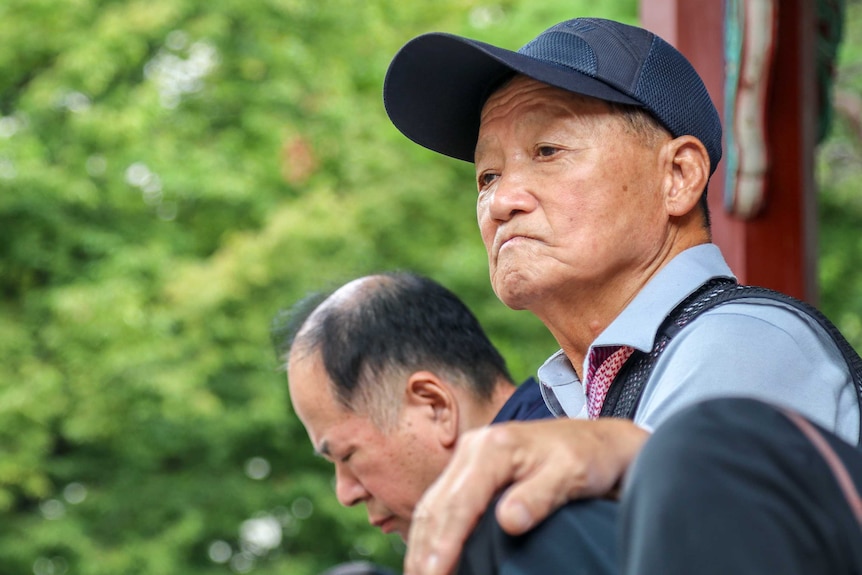 A elderly man in a baseball camps looks off frame against an unfocused leafy background
