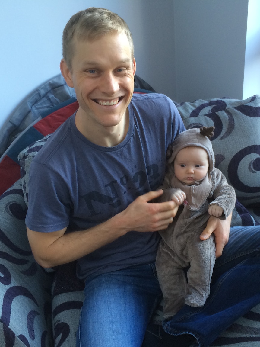 Un hombre rubio con una gran sonrisa cálida que sostiene a un pequeño bebé que lleva un pequeño mono con cuernos. 