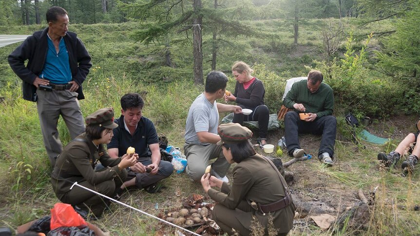 Foreigners and two women dressed in traditional North Korean uniform gather around a camp site.