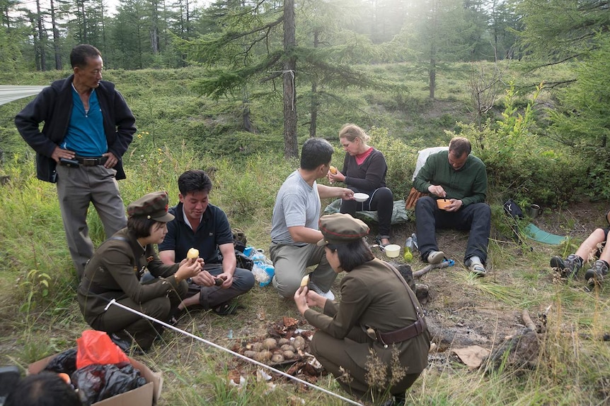 Foreigners and two women dressed in traditional North Korean uniform gather around a camp site.