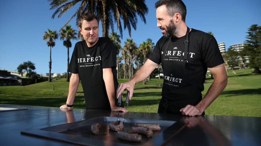 Two men in dark shirts chat while they tend to a barbecue in a park on a glorious day.