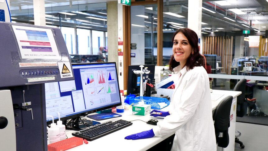 A woman in a lab coat smiles as she looks over her shoulder away from a computer