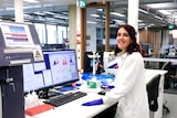 A woman in a lab coat smiles as she looks over her shoulder away from a computer