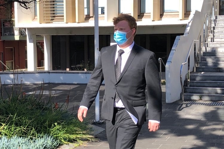 A ginger-haired man wearing a mask and a dark suit walks away from a court building.