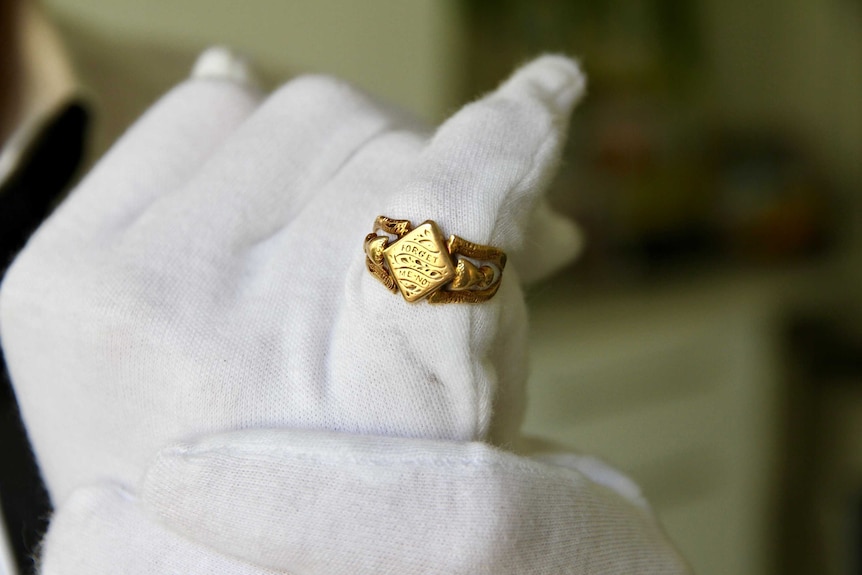 A white-gloved hand holding a gold ring, which says 'forget me not'