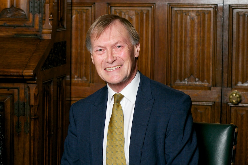 Photo shows David Amess sitting at a desk smiling at the camera 