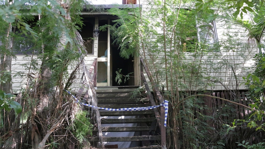 Police tape covers stairs of a high-set, cream wooden home, shrouded in plants.