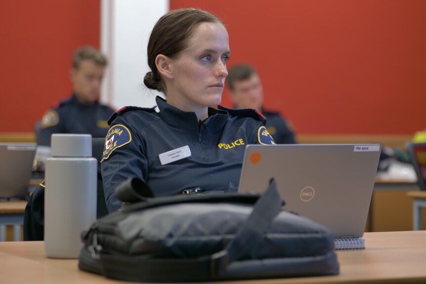 Young female police recruit in a classroom with a laptop and bag in front of her on a table.