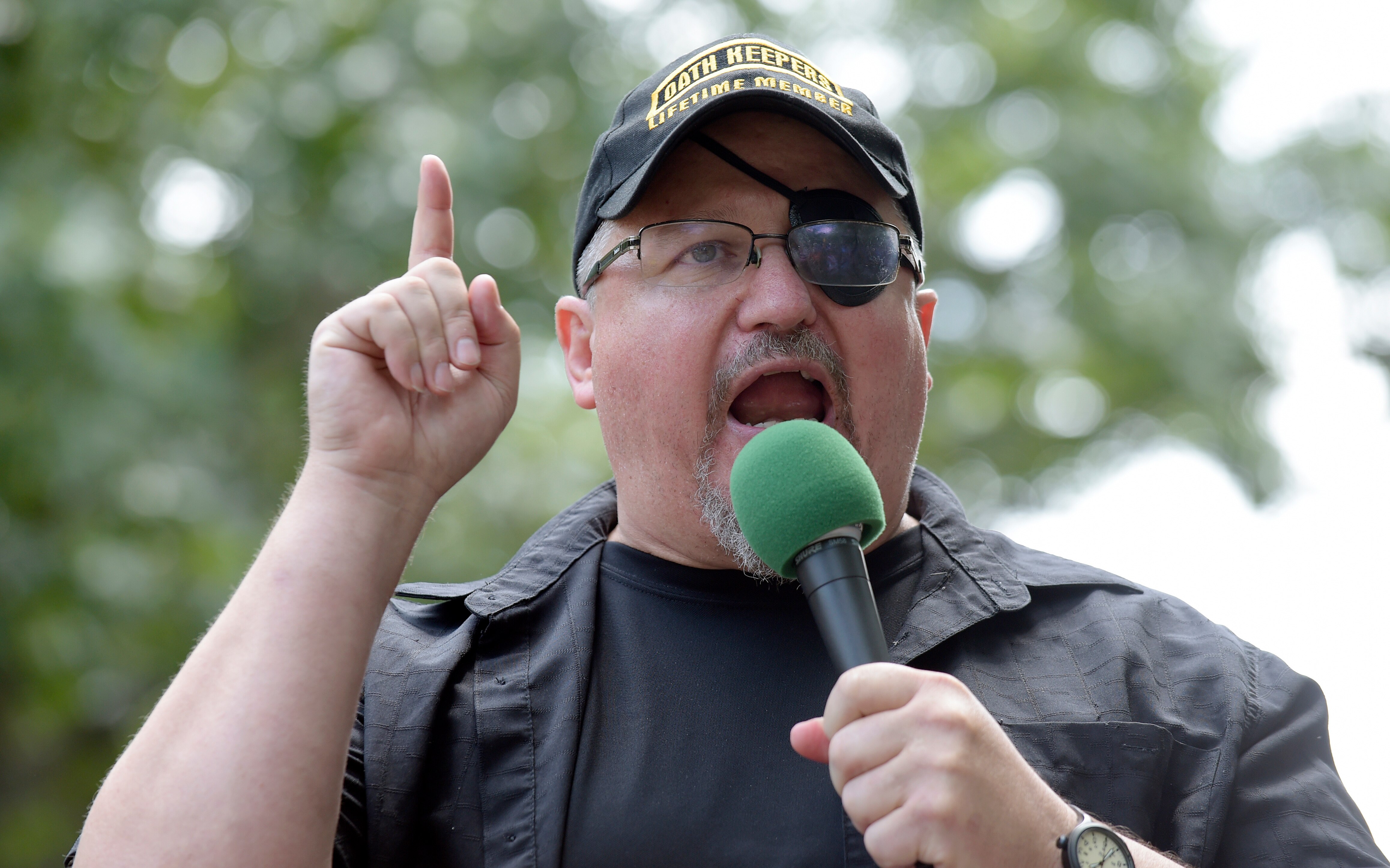A middle aged man is speaking into a microphone. He is dressed in black with a black eyepatch. He sticks one finger up.