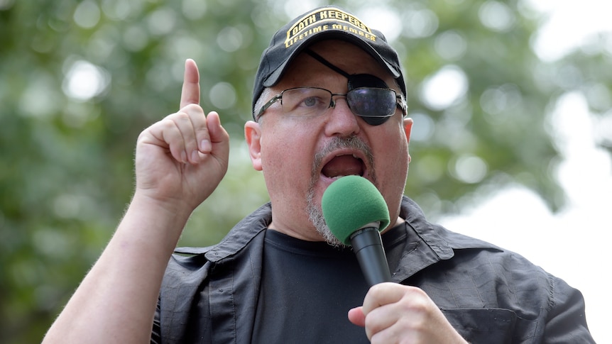 A middle aged man is speaking into a microphone. He is dressed in black with a black eyepatch. He sticks one finger up.