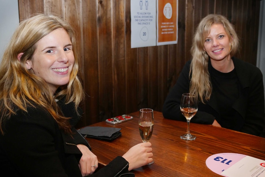 Two young women smile while sitting at a table with glasses of wine