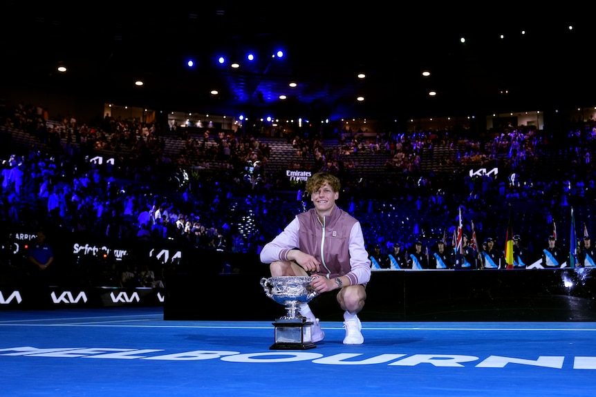 Tennis star Jannik Sinner crouches down on court to pose behind a big trophy after the Australian Open men's singles final.