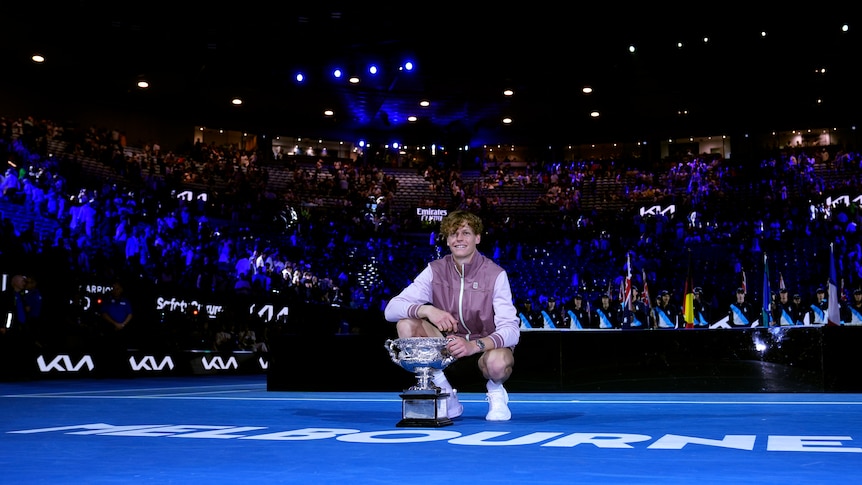 Tennis star Jannik Sinner crouches down on court to pose behind a big trophy after the Australian Open men's singles final.