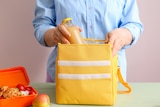 A woman puts a bottle of juice into an insulated yellow lunch bag ,a packed lunchbox sits beside it. Bringing lunch to work.