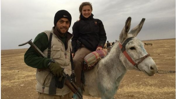 A teen girl sits on a donkey next to a man holding a rifle.