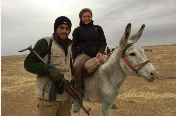 A teen girl sits on a donkey next to a man holding a rifle.