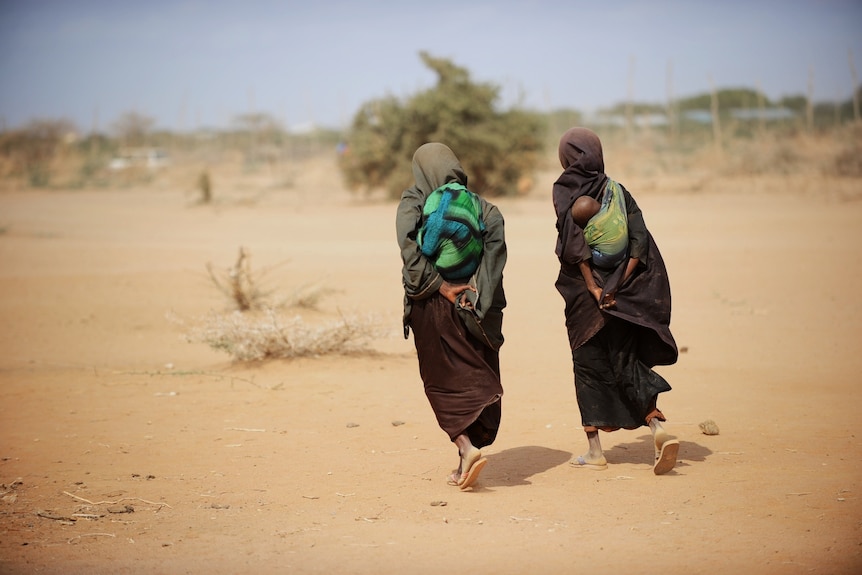 Two women, dressed all in black, with babies on their backs, walk through a sandy landscape in Africa