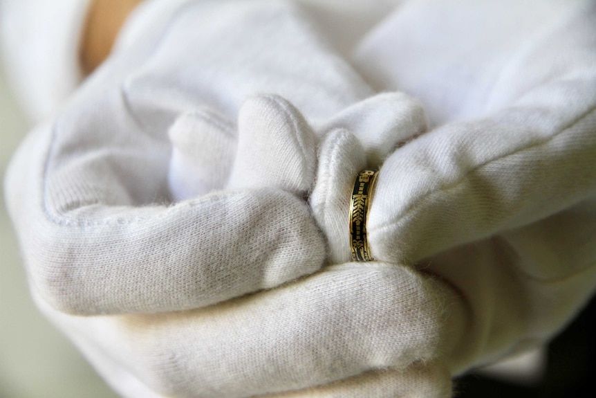 White-gloved hands holding a black enamel ring with a human skeleton around the band