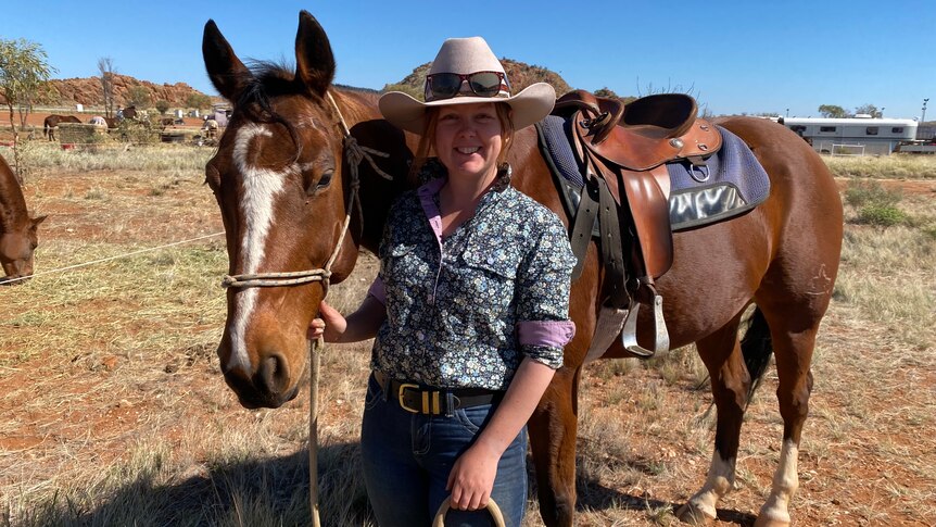 A young woman wearing a floral collared shirt, cowgirl hat and sunglasses stands at the shoulder of a bay horse in the outback.