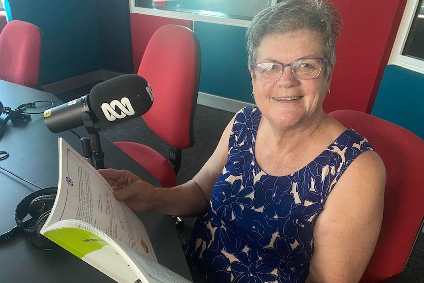 An elderly woman sits in a radio studio holding a cook book