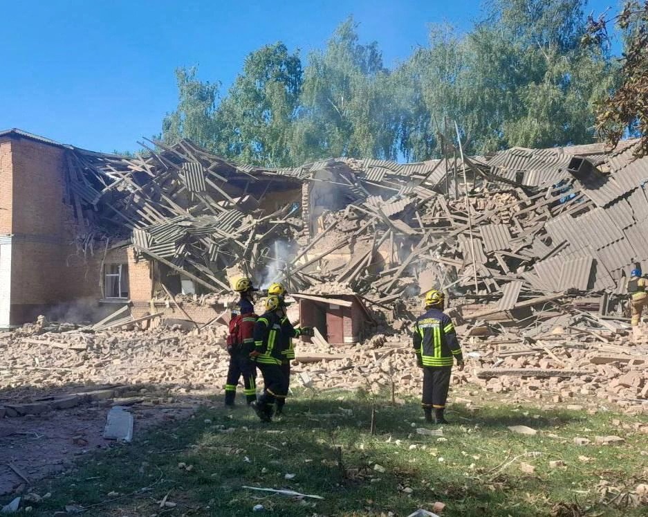 A collapsed building with bricks everywhere and trees behind it and four rescuers in front.