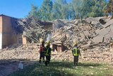 A collapsed building with bricks everywhere and trees behind it and four rescuers in front.