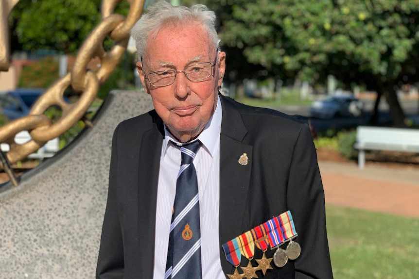 Elderly man stands wearing suit with army medals pinned to his chest and glasses on