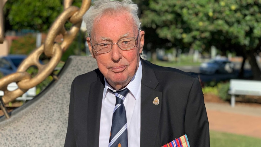 Elderly man stands wearing suit with army medals pinned to his chest and glasses on