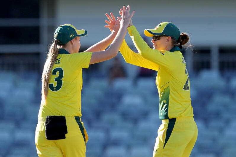 Two women in yellow and green cricket uniforms and caps high five each other on a cricket pitch