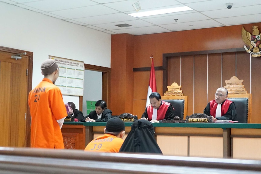 A man wearing an orange prison uniform stands in front of several judges in court robes