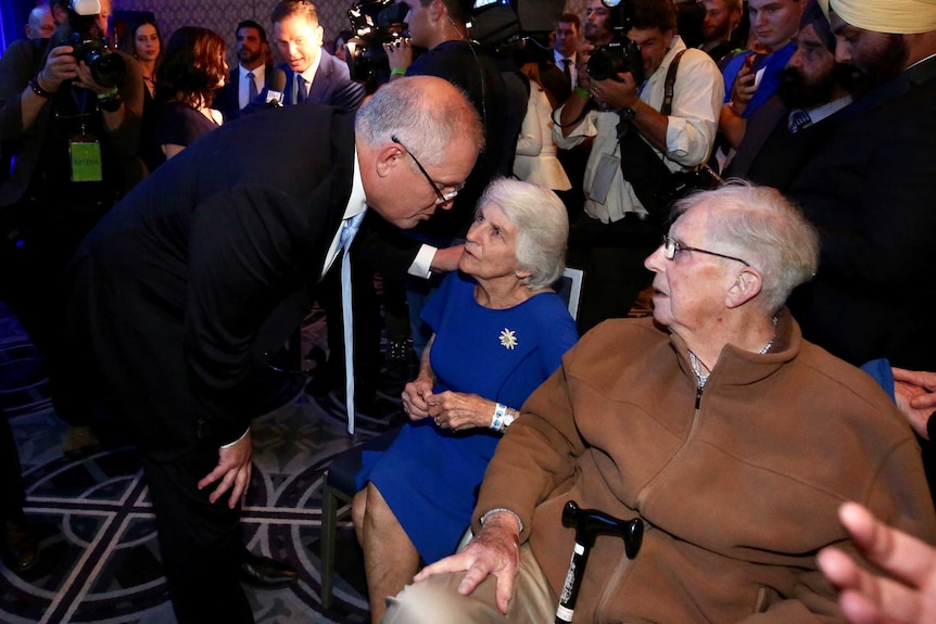 Scott Morrison puts his hand on his mother's shoulder as both his parents look up to him from seats