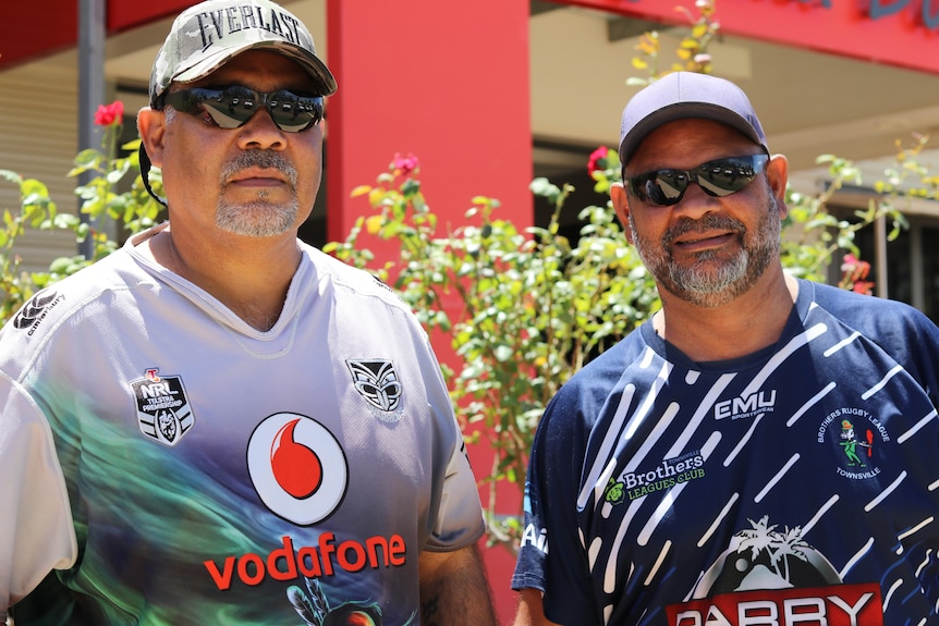 Two Waanyi Garawa men wearing multi-coloured shirts and caps stand next to eachother