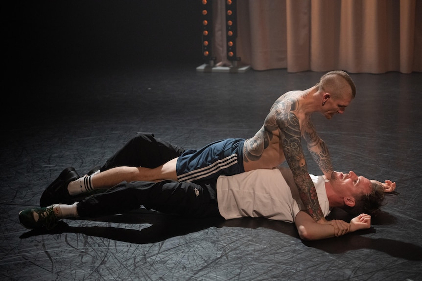 A shirtless man with tattoos lies atop of another man on a stage, pinning the man's arms behind his head.