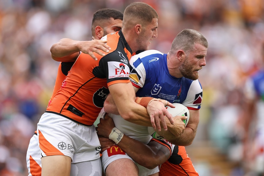 A Newcastle Knights NRL player holds the ball while being held by two Wests Tigers opponents.