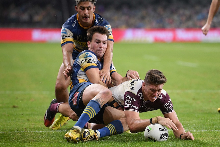 A Manly NRL player grounds the ball across the try line while being tackled by two Parramatta players.