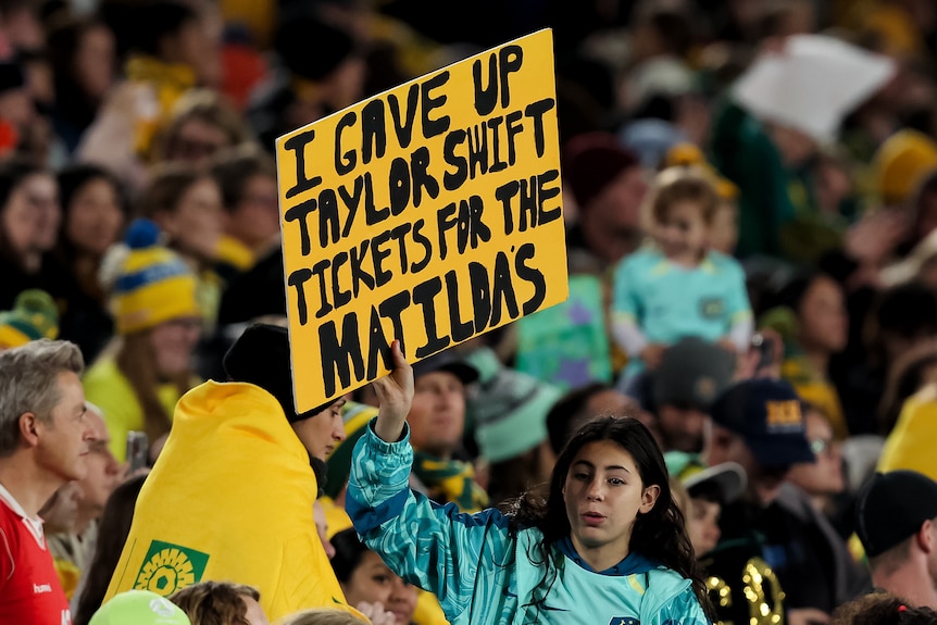 A young girl holds up a sign reading "I gave up Taylor Swift tickets for the Matildas" in the crowd of a Women's World Cup game.