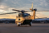 An army helicopter sits on the tarmac as the setting sun casts golden light across it from the left.