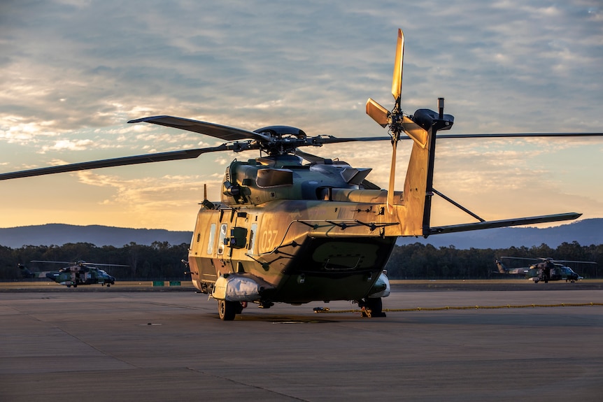 Un hélicoptère de l'armée est assis sur le tarmac alors que le soleil couchant projette une lumière dorée sur celui-ci depuis la gauche.