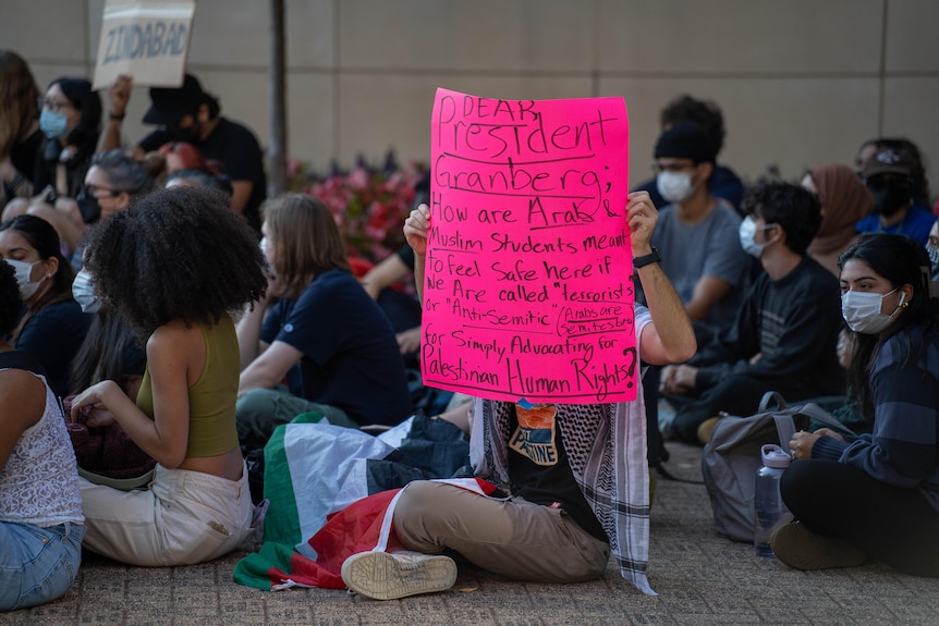 Students sit cross-legged on the ground. One holds a bright pink sign with pro-Palestinian messages.
