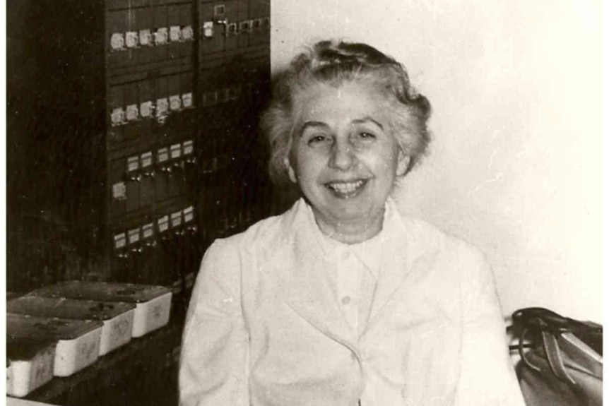 A black and white photo of a smiling woman sitting behind a desk