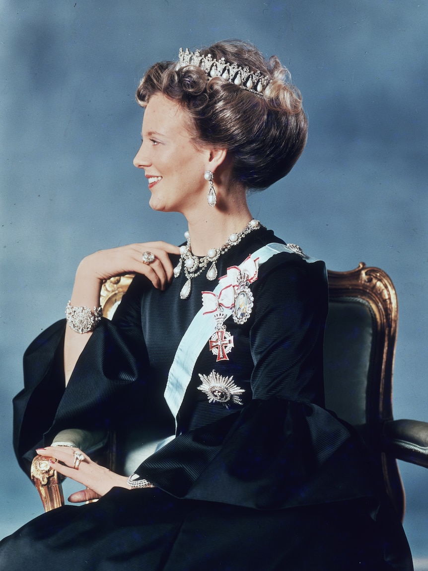 Queen Margrethe sits in a chair with crown and sash adorned with ornate jewellery