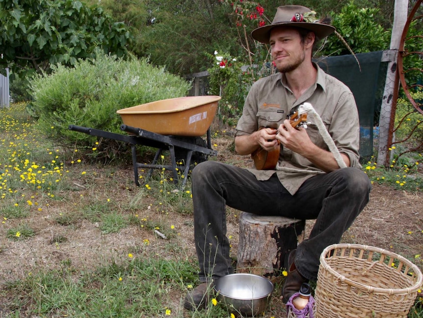 A man sitting on a tree stump playing the ukulele in a garden.