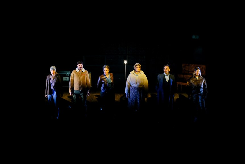 Six actors stand in a line onstage in various winter attire. They are spotlit and mid performance of the play Cyrano.