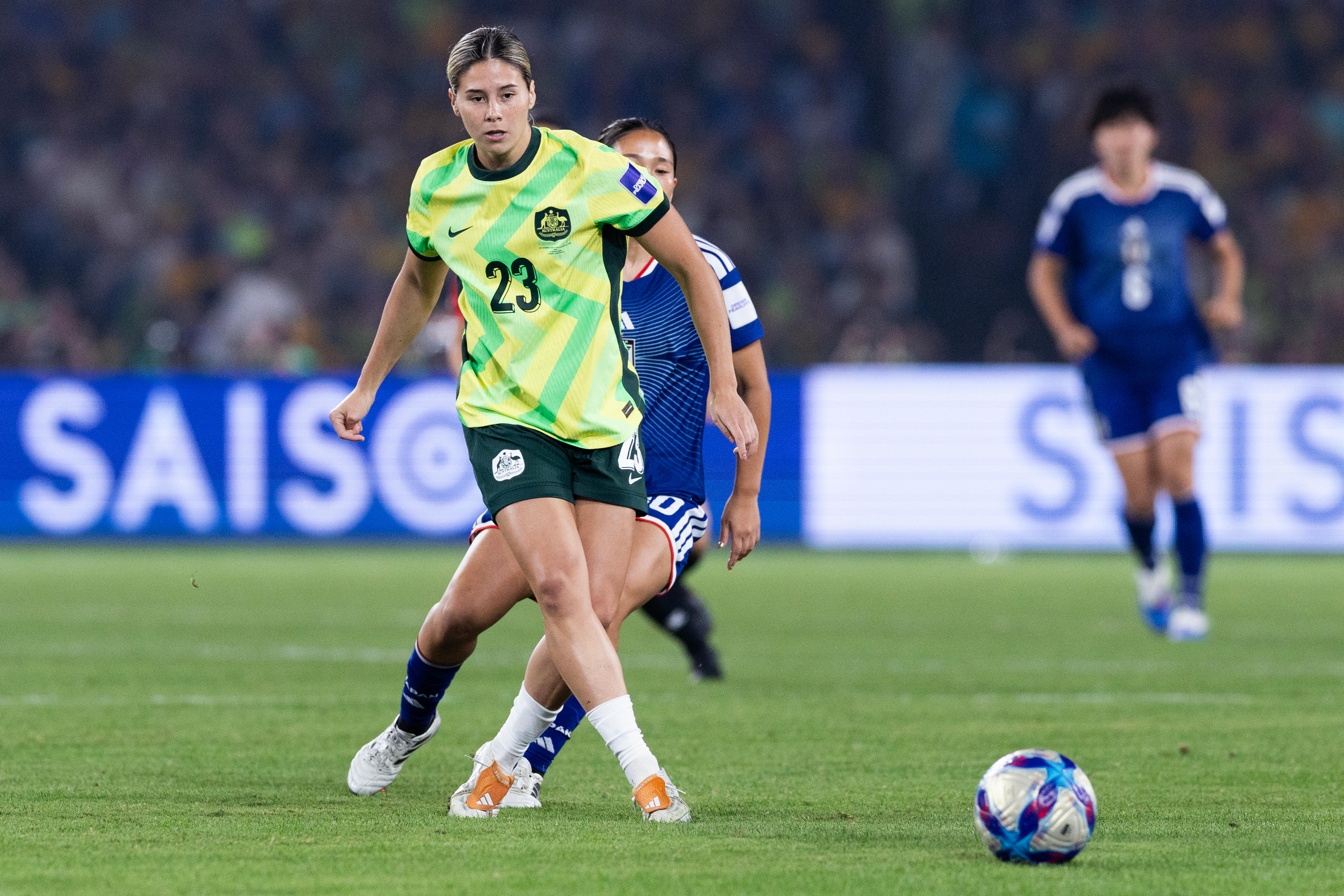 A soccer player in green and gold passes the ball with players in blue behind her