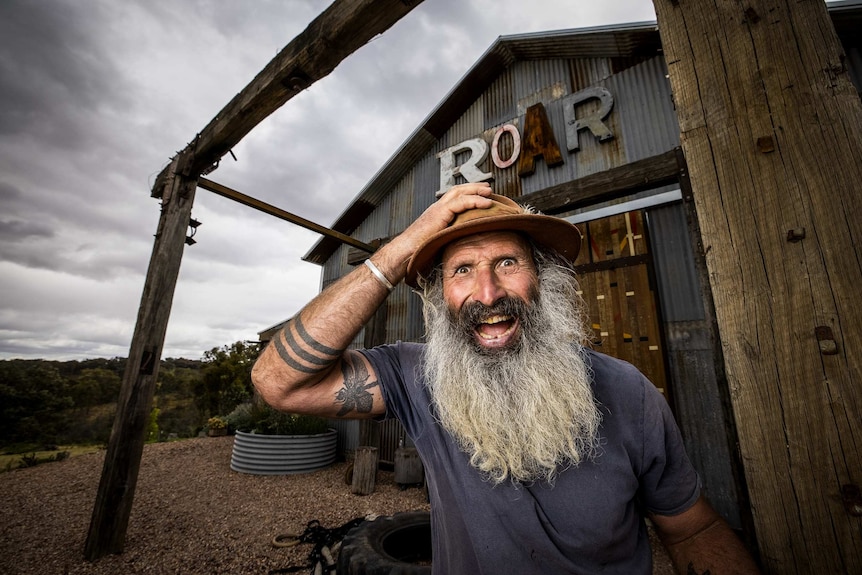 A man with a wild grey beard clutching a hat and looking jubilant