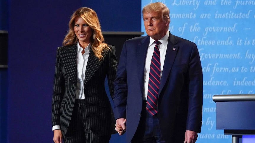 A woman in a pinstripe suit holds hands with a man in a navy suit while the holds hands and stand on a stage