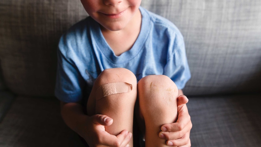 A young boy in a blue t-shirt smiles, holding his knees, one of which has a Band-Aid.