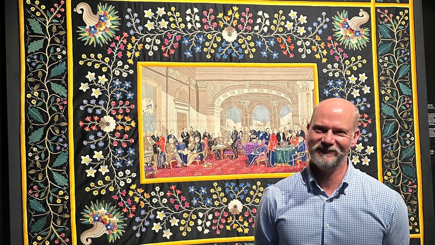 bald man with grey beard smiling in front of ornate historic quilt on a wall in full colour