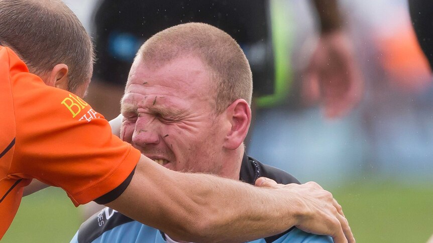 Luke Lewis grimaces with his eyes closed as a trainer (in orange) holds his shoulder.