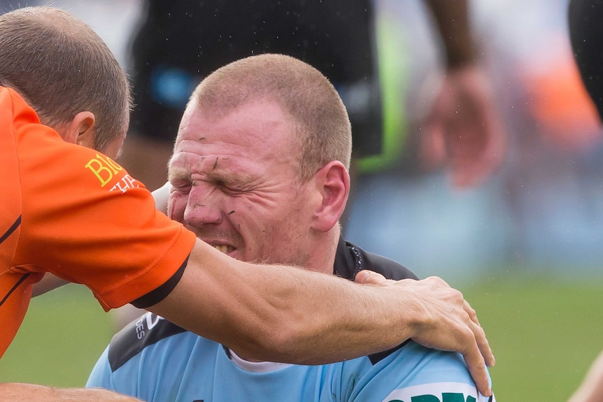 Luke Lewis grimaces with his eyes closed as a trainer (in orange) holds his shoulder.